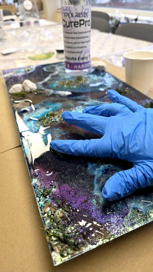 A hand wearing a blue nitrile glove smooths a glossy, colorful resin art board with a galaxy design. A bottle of Apex Resin CurePro resin is visible in the background on a table with other crafting supplies.
