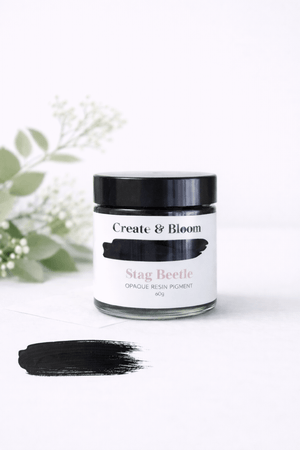 A jar of Create & Bloom Stag Beetle opaque resin pigment with a black lid and white label, shown on a white background with a black pigment swatch in front and soft green leaves blurred in the background.