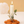 A white taper candle with a flame, styled in a flower-shaped terracotta holder with dried botanicals and green leaves, set against a warm beige background.