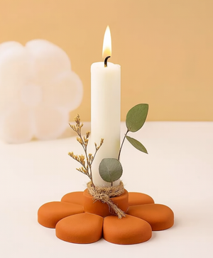 A white taper candle with a flame, styled in a flower-shaped terracotta holder with dried botanicals and green leaves, set against a warm beige background.