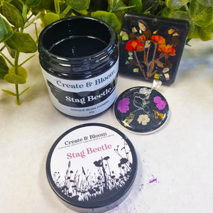 A jar of Create & Bloom Stag Beetle opaque resin pigment next to a square resin tile and a round resin coaster, all decorated with colorful pressed flowers and beetles, on a light surface with green leaves in the background.