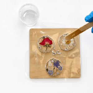 Three round resin pendants with pressed flowers are arranged on a wooden board, with a gloved hand using a wooden stick to spread liquid resin over one pendant. A small clear cup is visible at the top left on a white background.