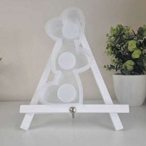 A white wooden tabletop easel stand with a small hanging silver key or clasp at the center, shown on a light surface with a potted green plant in the background.