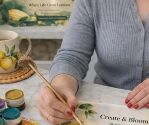 Person holding a paintbrush with art supplies and a book titled 'Create & Bloom' on a table.