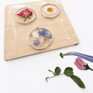Three round silver-toned pendants with pressed flowers and green leaves, displayed on a wooden board with clear tape. A pair of tweezers holds a pink flower bud below on a white background.